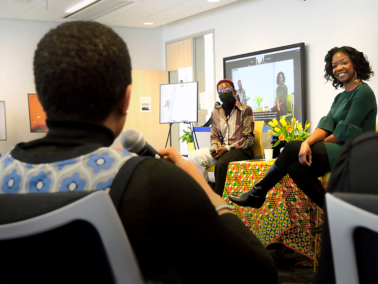 Two people sat next to each other in front of a person speaking into a microphone. 